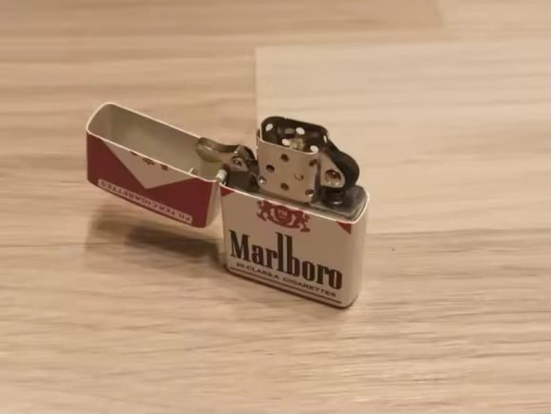 Puede incluir: Un encendedor de metal con tapa abatible en acabado blanco y rojo que presenta el logotipo y el texto de Marlboro. El encendedor est&aacute; abierto, revelando la chimenea interna plateada y el mecanismo de la rueda. El exterior muestra la marca y el texto 20 Class A Cigarettes.