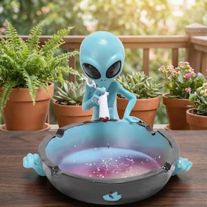 Puede incluir: Una figura de alienígena azul claro sosteniendo un cigarrillo, encima de un cenicero negro y azul. El cenicero tiene un interior con temática de galaxia. La figura y el cenicero están sobre una superficie de madera, con plantas en macetas en el fondo.