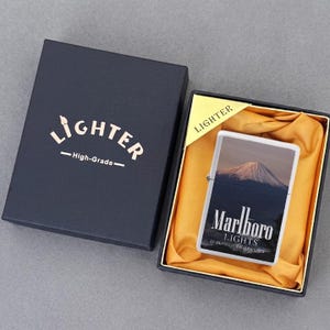 Puede incluir: Un encendedor met&aacute;lico rectangular con una imagen del monte Fuji y el texto Marlboro LIGHTS 20 CLASS A CIGARETTES, presentado en una caja de regalo negra con forro de tela dorada y el texto LIGHTER High-Grade impreso en la tapa.