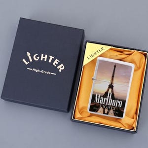 Puede incluir: Un encendedor de metal rectangular con una imagen de la Torre Eiffel al atardecer y el texto Marlboro LIGHTS, presentado en una caja de regalo azul oscuro con forro de tela dorada y la palabra LIGHTER impresa en la tapa y en un inserto.