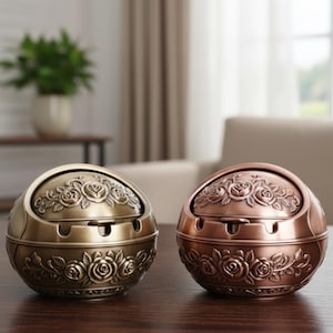 May include: Two decorative metal ashtrays. One is bronze-colored, the other is copper-colored. Both are round with a hinged lid and rose floral design. The ashtrays are sitting on a dark wooden surface.