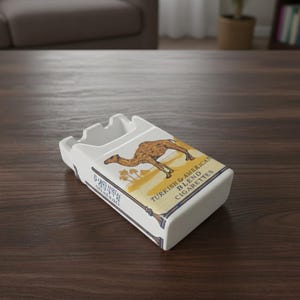 May include: White ceramic ashtray in the shape of a cigarette pack, with a camel illustration and the text "TURKISH & AMERICAN BLEND CIGARETTES". The ashtray is rectangular with grooves for cigarettes.