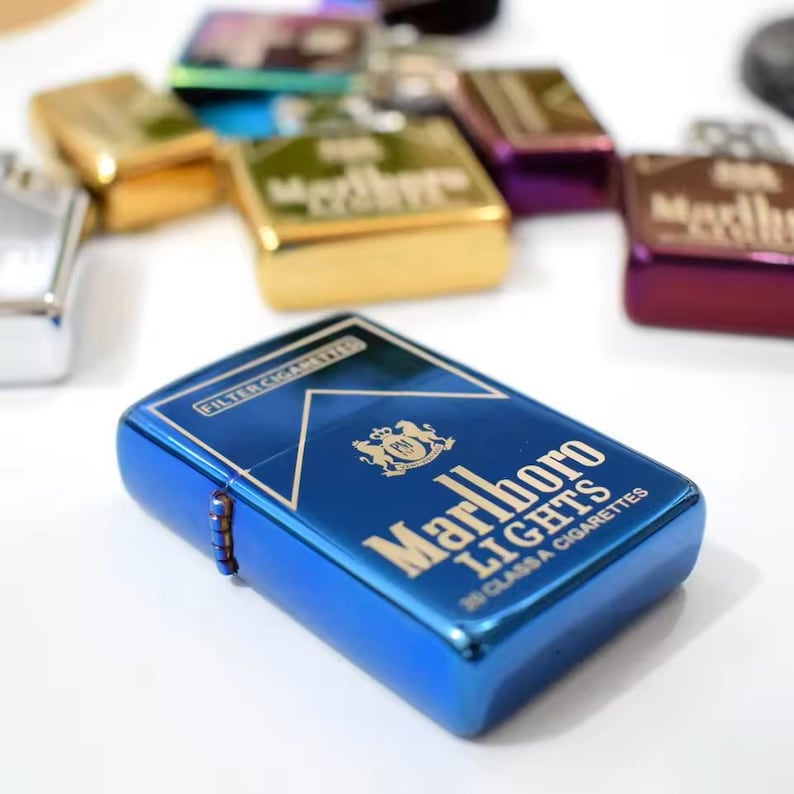 Puede incluir: Un encendedor met&aacute;lico azul con tapa abatible que presenta el logotipo de Marlboro Lights en dorado, incluyendo el texto Filter Cigarettes y 20 Class A Cigarettes. Se ven otros encendedores en varios colores met&aacute;licos como oro, morado y plata.