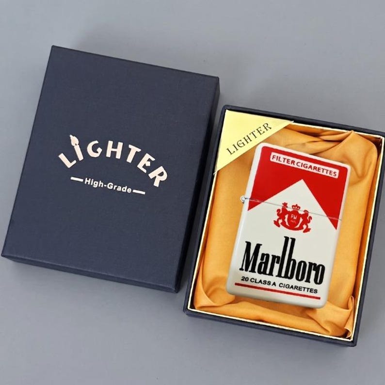 Puede incluir: Un encendedor met&aacute;lico rectangular con un dise&ntilde;o en rojo y blanco que incluye el logotipo y texto de Marlboro, presentado en una caja de regalo azul oscuro con forro de tela dorada. La tapa muestra la palabra LIGHTER y High-Grade.