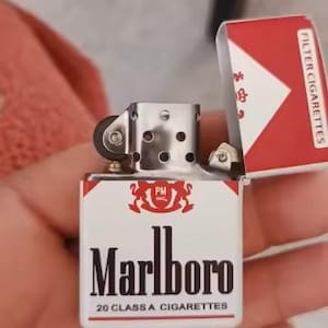 Puede incluir: Un encendedor de metal con tapa abatible que presenta un dise&ntilde;o en blanco y rojo con el logotipo de Marlboro, el texto PM Inc. y las palabras 20 Class A Cigarettes. La tapa est&aacute; abierta, revelando el mecanismo interno plateado con una rueda negra.