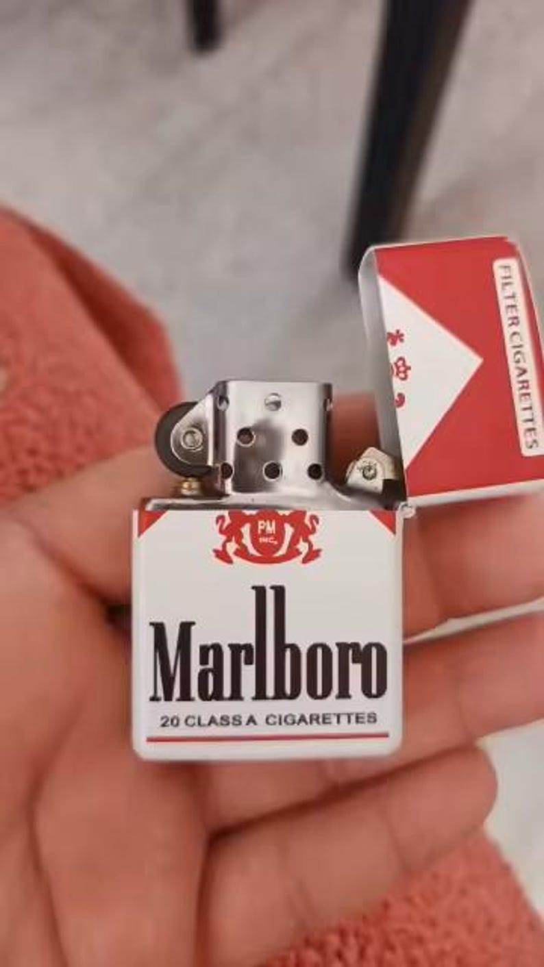 Puede incluir: Un encendedor de metal con tapa abatible que presenta un dise&ntilde;o en blanco y rojo con el logotipo de Marlboro, el texto PM Inc. y las palabras 20 Class A Cigarettes. La tapa est&aacute; abierta, revelando el mecanismo interno plateado con una rueda negra.
