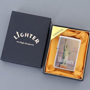 Puede incluir: Un encendedor met&aacute;lico rectangular con una imagen de la Estatua de la Libertad y el texto Marlboro Lights, presentado en una caja negra con forro de tela dorada. La tapa negra tiene el texto LIGHTER High-Grade impreso en color dorado.