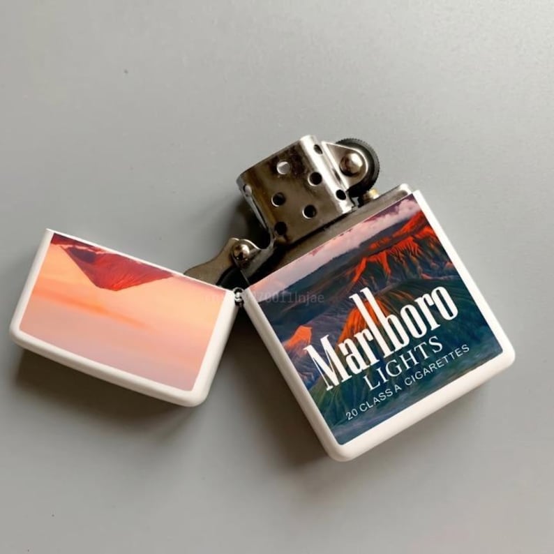 Puede incluir: Un encendedor de metal con tapa abatible y carcasa blanca que presenta un gr&aacute;fico de monta&ntilde;as al atardecer. El texto Marlboro LIGHTS 20 CLASS A CIGARETTES est&aacute; impreso en el frente. La bisagra de metal plateado es visible.