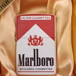 Puede incluir: Un encendedor met&aacute;lico rectangular con la marca de cigarrillos Marlboro en rojo y blanco, incluyendo el texto Filter Cigarettes, el escudo PM Inc., Marlboro y 20 Class A Cigarettes, presentado dentro de una caja forrada en color dorado.
