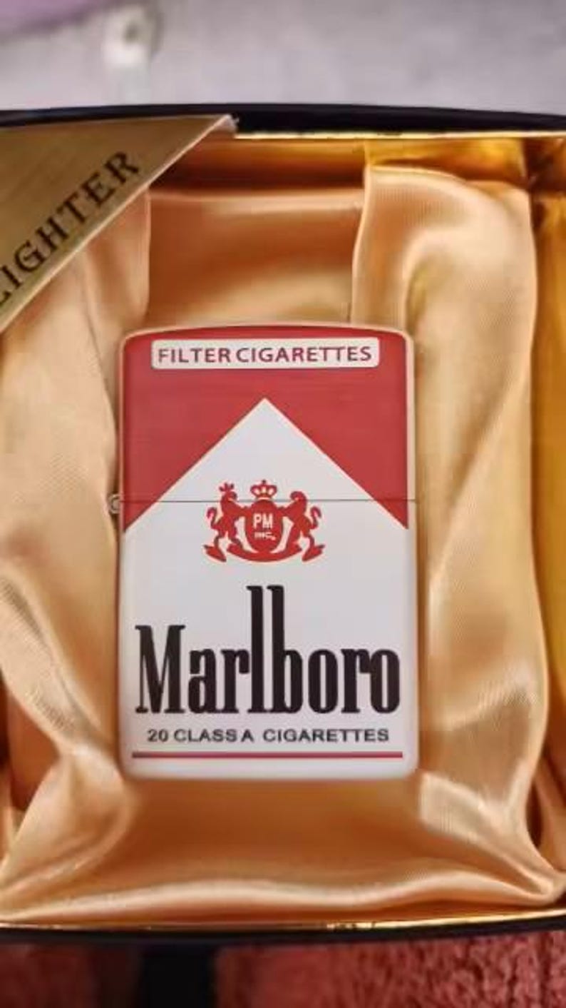 Puede incluir: Un encendedor met&aacute;lico rectangular con la marca de cigarrillos Marlboro en rojo y blanco, incluyendo el texto Filter Cigarettes, el escudo PM Inc., Marlboro y 20 Class A Cigarettes, presentado dentro de una caja forrada en color dorado.