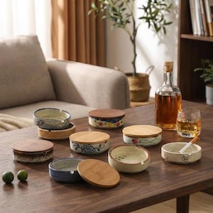 以下が含まれることがあります： 木製の蓋が付いた、花柄や模様が施された陶器の灰皿のコレクション。灰皿は青、ベージュ、茶色など様々な色があります。琥珀色の液体が入ったボトルとウイスキーのグラスもテーブルに置かれています。