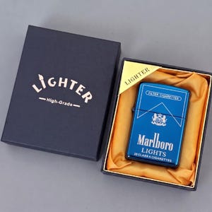 Puede incluir: Un encendedor de metal azul con tapa abatible que presenta el logotipo y el texto de Marlboro Lights, presentado en una caja de regalo azul oscuro con forro de tela dorada. La tapa de la caja muestra la palabra LIGHTER y High-Grade en blanco.