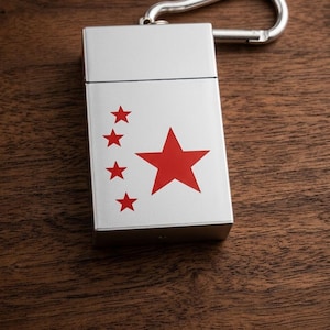 May include: A silver metal cigarette case with a red star and four smaller red stars. The case has a hinged lid and a silver carabiner clip attached to the top. The case is rectangular and sits on a wooden surface.