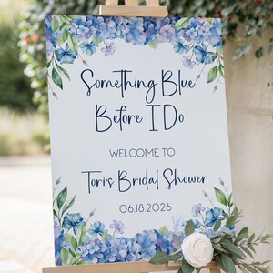 May include: A wooden easel displays a sign with the text "Something Blue Before I Do" and "Welcome to Tori's Bridal Shower 06.18.2026". The sign is decorated with blue and purple floral accents and a white rose.