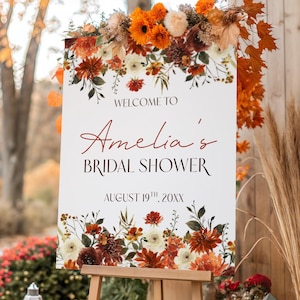 Może przedstawiać: Prostokątny biały znak na drewnianej sztaludze z napisem "WELCOME TO Amelia's BRIDAL SHOWER AUGUST 19TH, 20XX" czcionką w kolorze czarnym i czerwonym, ozdobiony kompozycją kwiatową z pomarańczowych, czerwonych i białych kwiatów.
