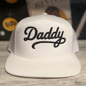 Op de afbeelding: Witte baseballcap met een platte klep en mesh achterkant. Het woord "Daddy" is in zwart script op de voorkant geborduurd. De cap heeft een klassiek trucker-hoed ontwerp.