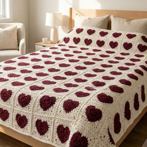 May include: A cream-colored, crocheted blanket with a repeating pattern of burgundy hearts. The blanket is draped over a wooden bed frame, with a matching cream-colored chair and side table in the background. The blanket is approximately 80 inches by 90 inches.