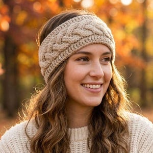 May include: A beige cable-knit headband is worn by a person with long, wavy brown hair. The headband is made of a soft, textured yarn. The person is smiling, and the background is a blurred autumn scene with trees and falling leaves.