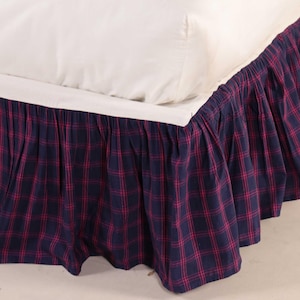 Puede incluir: Una falda de cama a cuadros azul marino y rojo con un diseño plisado. La falda de cama está hecha de una tela suave y está diseñada para encajar alrededor de la base de una cama. La sábana y la almohada blancas son visibles.