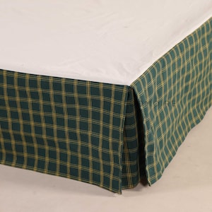 Może przedstawiać: Spódnica do łóżka z zielono-żółtym wzorem w kratę. Tkanina jest udrapowana wokół podstawy łóżka, z białym prześcieradłem widocznym na górze. Wzór w kratę przedstawia siatkę żółtych linii na ciemnozielonym tle.