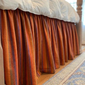 May include: A bed skirt with vertical stripes in shades of orange, purple, and brown. The skirt is draped under a white comforter, creating a layered look. The fabric has a textured appearance, adding depth to the design.