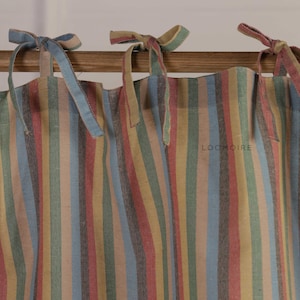 May include: A curtain featuring vertical stripes in muted tones of blue, green, yellow, and red. The fabric is suspended from a wooden rod by tied fabric loops at the top. The text LOOMOIRE is printed in small, dark letters on the fabric surface.