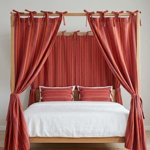 May include: A wooden canopy bed with red and orange striped curtains tied back with matching fabric. Two striped pillows rest on the bed, which is covered with a white comforter. The bed frame is a light wood color.