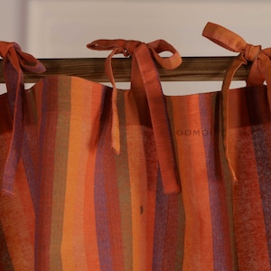 May include: A curtain featuring vertical stripes in shades of orange, red, purple, and green. The fabric is suspended from a wooden rod by several tied fabric ribbons at the top. The word LOOMOIRE is printed in small, faint letters on the fabric.