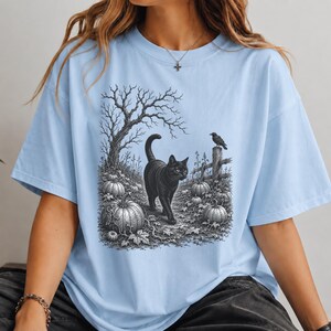 May include: Light blue t-shirt featuring a black and white illustration of a black cat walking through a pumpkin patch. A crow sits on a fence post near a bare tree. The design has a vintage, Halloween theme.