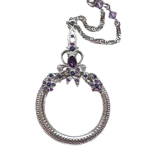 May include: A silver-toned magnifying glass pendant on a matching chain. The circular frame features intricate metalwork, accented with small purple and dark blue stones. A larger oval purple stone sits at the top, with additional purple beads on the chain.