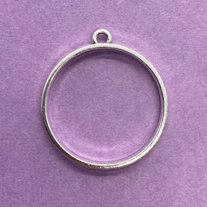 May include: A silver-colored metal pendant frame with a circular design. The frame has a small loop at the top for attaching to a chain or other jewelry component. The pendant is set against a purple background.