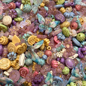 May include: A collection of assorted craft beads in various shapes and sizes, including butterfly-shaped beads, floral motifs, and textured spheres in a palette of pastel pink, lavender, mint green, cream, and iridescent blue with gold accents.