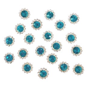 May include: A collection of turquoise-colored rhinestone embellishments. Each rhinestone is round with a central turquoise stone surrounded by a ring of smaller, clear rhinestones. The embellishments are scattered on a white background.