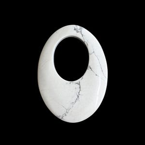 May include: White oval pendant with a central opening, featuring gray veining throughout. The polished stone is set against a black background, highlighting its smooth texture and natural patterns. A unique jewelry piece.