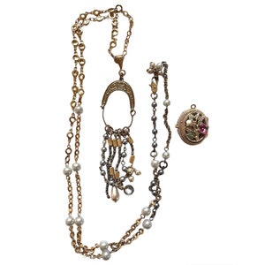 May include: A gold necklace with a long pendant featuring a horseshoe design and a tassel of gold and silver beads. The necklace also has a smaller pendant with a floral design and a pink flower.