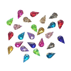 May include: A collection of teardrop-shaped rhinestones in various colors, including pink, red, blue, green, and gold. Each gem has a metallic gold setting. The rhinestones are scattered on a white background.