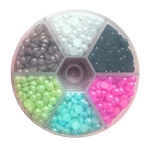 May include: A clear, round container divided into six sections, each filled with colorful, half-round faux pearls. Colors include gray, white, dark teal, pink, light green, and turquoise. The container has a central, hexagonal hub.