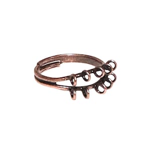 May include: A bronze-colored ring with a double band design. The top band features a series of small, circular loops, while the lower band has three vertical, rounded accents. The ring is set against a white background.