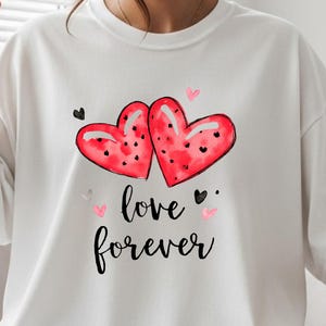 Könnte beinhalten: Weißes T-Shirt mit einem Aquarelldesign aus zwei roten Herzen mit schwarzen Tupfen und den Worten "love forever" in schwarzer Schrift. Das Design enthält kleine schwarze und rosa Herzen.