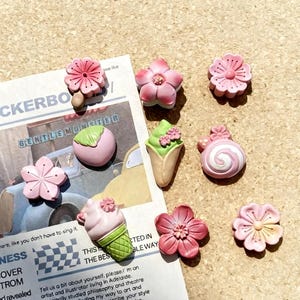 May include: A selection of pink and cream resin charms, including flower shapes, an ice cream cone, and a swirl. The charms are displayed on a newspaper with text.