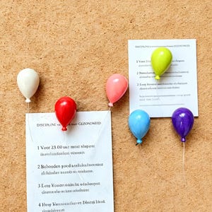 May include: A corkboard with six balloon-shaped push pins in various colours: white, red, pink, blue, green, and purple. Two white papers are pinned to the board with text. The push pins are used to hold the papers.