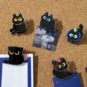 Puede incluir: Una colección de clips acrílicos con forma de gato negro con varios diseños. Algunos gatos tienen ojos amarillos o azules, y algunos llevan accesorios como un collar o un gorro de fiesta. Los clips están sujetos a papel y a un fondo de corcho.