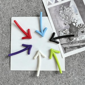 May include: A selection of colourful arrow-shaped magnets in red, blue, black, purple, white, and green. The magnets are arranged on a white square, with a black and white print in the background. These magnets are ideal for use on a fridge or a magnetic board.