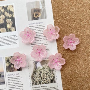 May include: Six translucent pink flower-shaped embellishments with glitter. Each flower has five petals and a darker pink centre. They are scattered on a printed paper background, suitable for crafting or jewellery making.