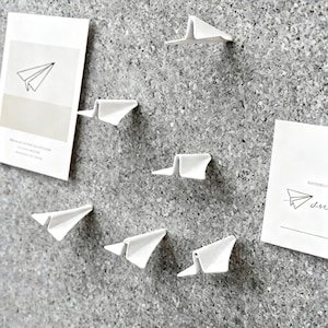 May include: White paper airplane-shaped push pins and two cards with a paper airplane graphic are attached to a grey corkboard. The push pins are arranged in a scattered pattern, creating a decorative and functional display.