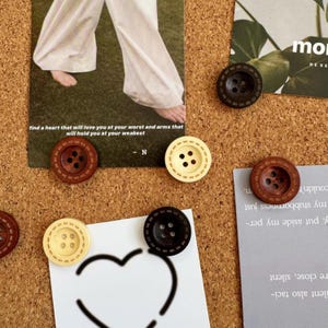 May include: A corkboard displaying decorative buttons in shades of brown, black, and cream. The buttons feature a stitched border and are used to hold up cards with text. One card reads "find a heart that will love you..."