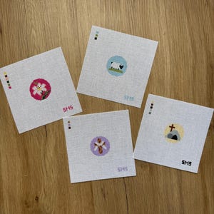 May include: Four square white needlepoint canvases featuring circular designs: a pink circle with a white flower, a blue circle with a sheep, a purple circle with a cross, and a yellow circle with a tomb and cross. Each canvas includes color keys and the text SMS.