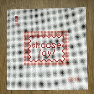May include: A white needlepoint canvas featuring a central red and pink checkered square border with a scalloped edge. Inside the border, the text "choose joy!" is stitched in red. A small red color palette and the letters "SMS" are visible.