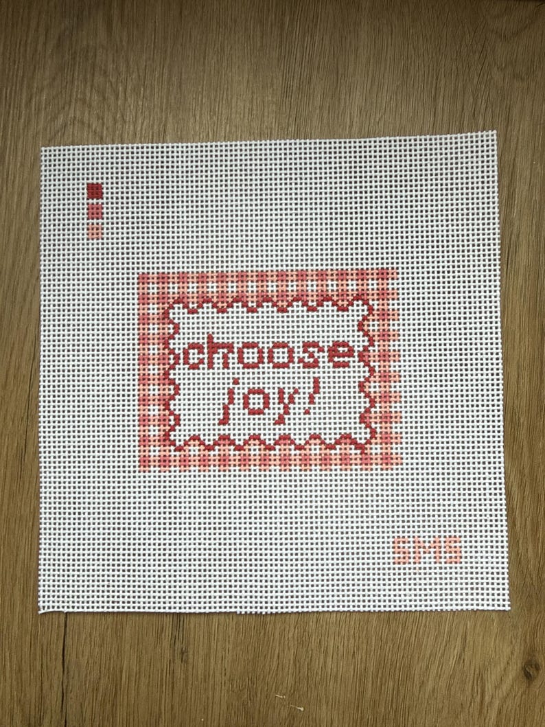 May include: A white needlepoint canvas featuring a central red and pink checkered square border with a scalloped edge. Inside the border, the text "choose joy!" is stitched in red. A small red color palette and the letters "SMS" are visible.