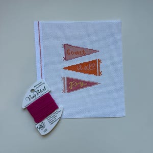 K&ouml;nnte beinhalten: Eine Gobelin-Leinwand mit drei Wimpeln, die in Orange- und Rosat&ouml;nen gestickt sind, jeweils mit einem Wort: "Count", "it all" und "joy". Eine Spule rosa Very Velvet-Garn befindet sich in der linken unteren Ecke.
